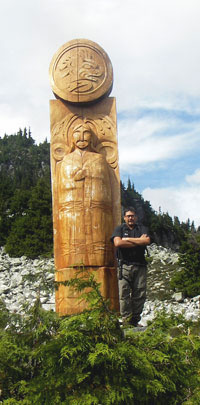 Man standing next to  large post carved with first nations designs.